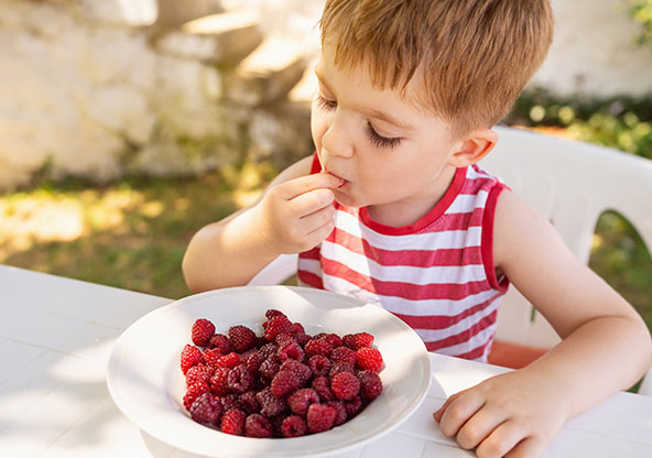A young boy is eating a bowl of raspberries at a table outside.