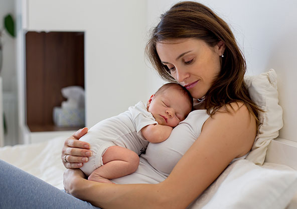 A new mom is sitting on her bed, holding her newborn baby who is sleeping on her chest