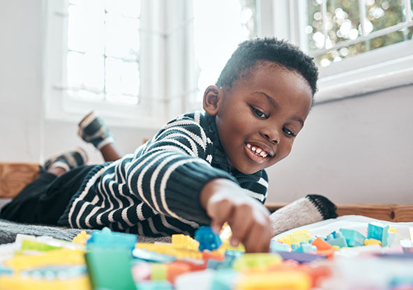 A young, happy boy is laying on the ground in his house playing with blocks by himself.