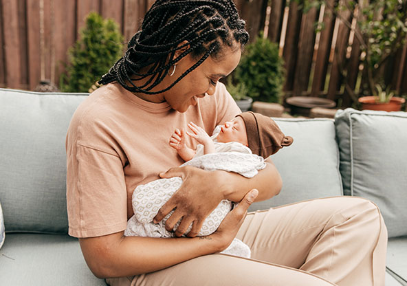 A young black mom is holding her newborn baby outside on a couch in the backyard.