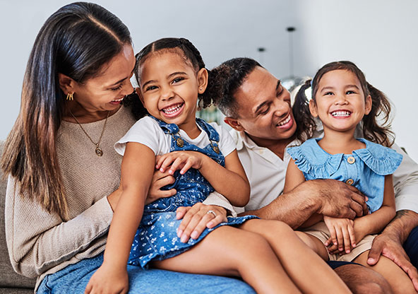 Two little girls and their parents are sitting on the couch together smiling