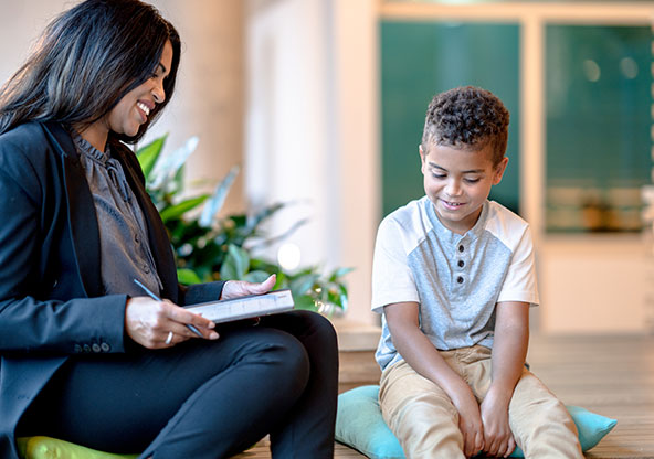 A young boy is sharing and talking with a child psychologist in therapy. She is smiling and taking notes while she listens to him.