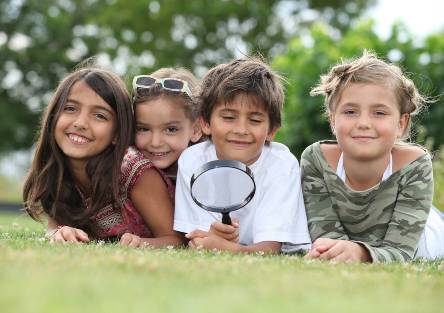 Several smiling children sit together in the grass, and one holds a magnifying glass