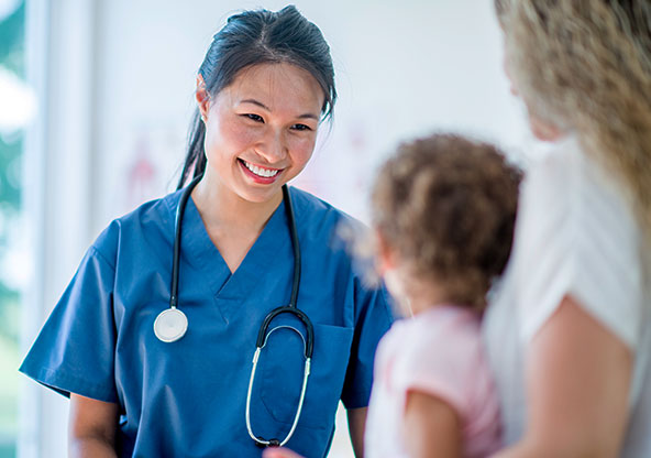 A nurse at an urgent care smiling at a little girl who is being held by her mom