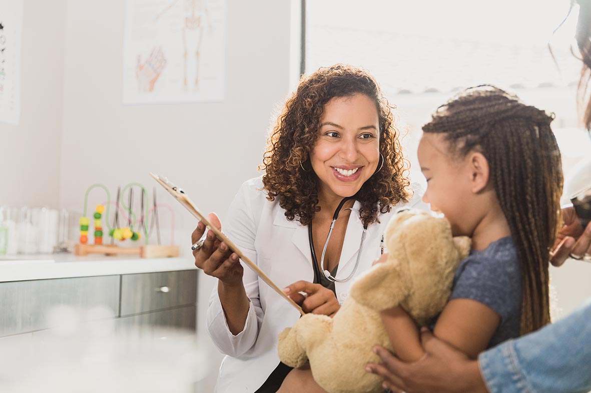 A preschool age girl holds her stuffed animal toy as she talks with a female pediatrician during a well visit.